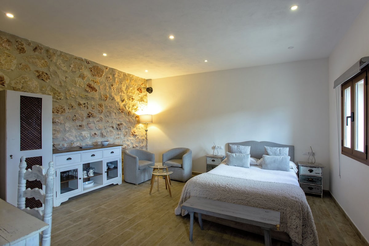 Une chambre avec un mur en pierre, un lit avec des oreillers, une table de chevet, une lampe, une chaise, une petite table et un meuble.