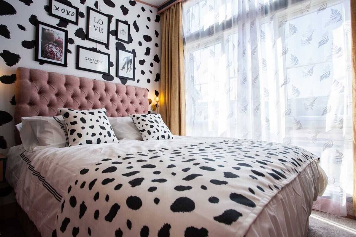 Quarto com cabeceira estofada, cama com colcha de manchas pretas e brancas e janela com cortinas translúcidas.