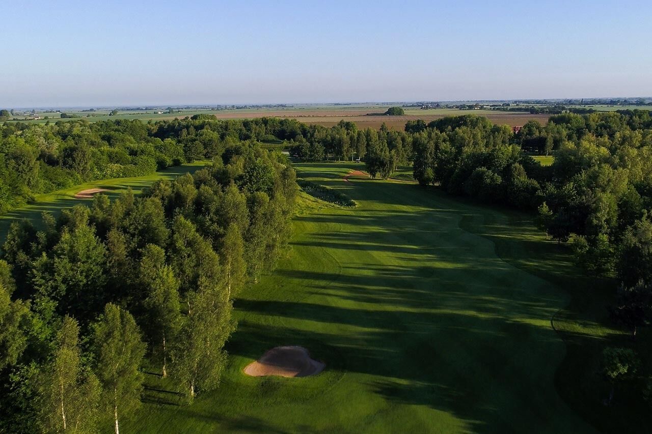 An aerial view of a golf course with a large forested area, a clear sky, and distant fields.