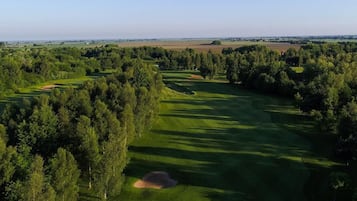 An aerial view of a golf course with a large forested area, a clear sky, and distant fields.