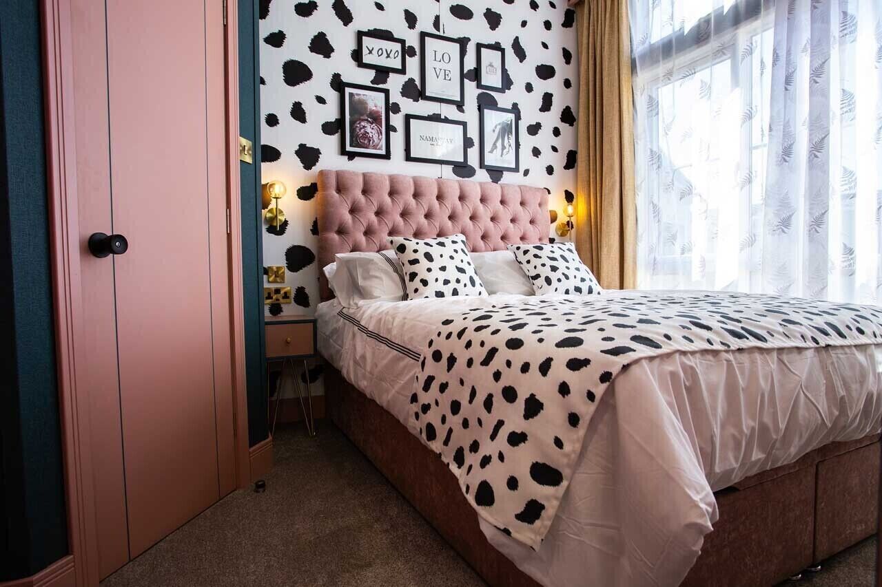 A bedroom with a tufted headboard, a bed with a white and black spotted blanket, a nightstand with a lamp, and a wall with framed pictures and a cow print pattern.