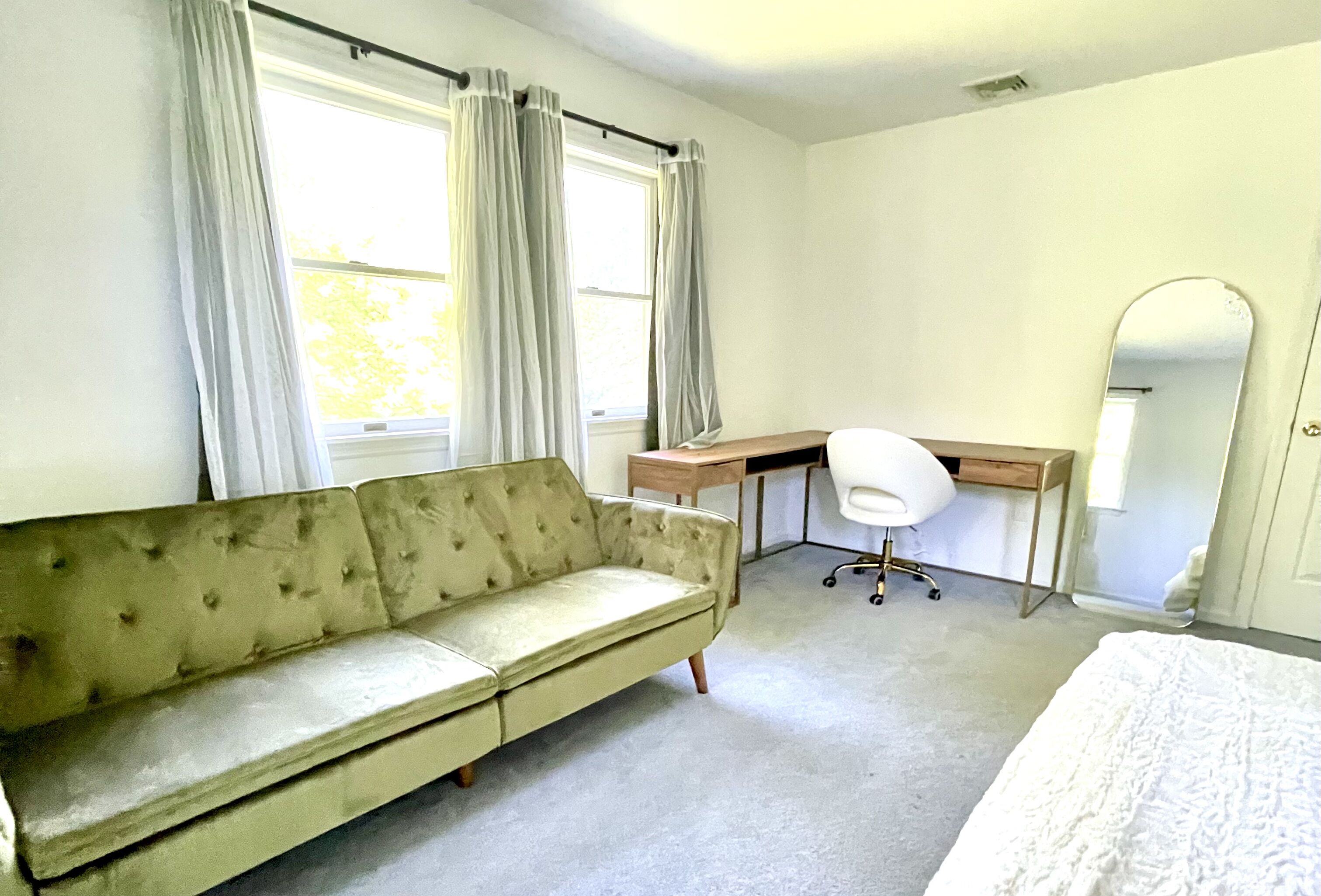 A room with a beige sofa, a wooden desk, a white chair, and a bed.