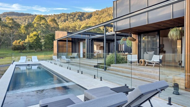 Ein moderner Poolbereich im Freien mit Liegestühlen, einer Holzterrasse und einer glasgeschlossenen Veranda mit Sitzgelegenheiten und einer hängenden Pflanze.