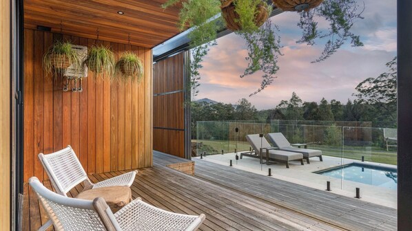 A modern outdoor deck with wooden flooring, two wicker lounge chairs, a glass-enclosed pool, and hanging plants.