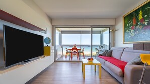 Uma sala de estar moderna com uma televisão de tela plana, um espaço de refeições com mesa e cadeiras, e um sofá com um cobertor vermelho.