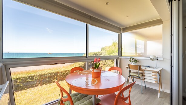 Uma área de refeições moderna com uma grande janela com vista para uma praia, uma mesa redonda laranja e cadeiras laranjas.