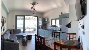 A modern living room with a sofa, dining table, and chairs, a kitchen with cabinets and appliances, and a large window providing natural light. - Pythagorion Harbour Residence (Pythagoreio)