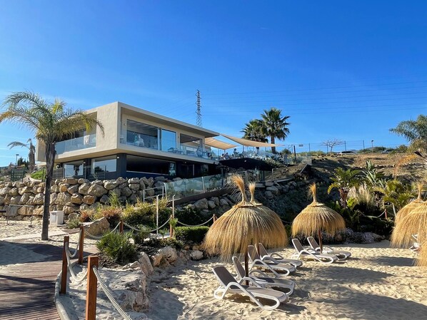 Ein modernes Strandhaus mit einem Balkon mit Blick auf den Sand, umgeben von Palmen und Reetdachschirmen.