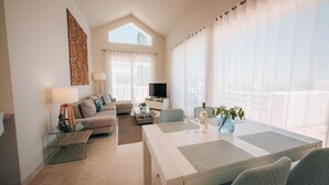 Un salon moderne avec un canapé blanc, une table basse en verre, un téléviseur et un coin repas comprenant une table et des chaises blanches.