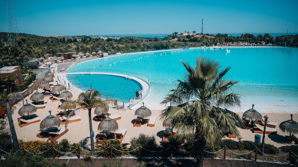 Un complexe hôtelier doté d’une grande piscine, d’une plage avec des chaises longues et des parasols, et offrant une vue sur l’océan.