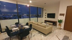 Una sala de estar moderna con comedor, ventanas amplias y vista a la ciudad, un sofá, televisor y planta.
