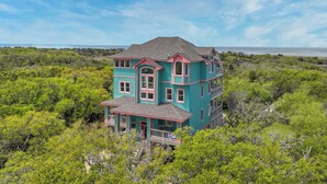 A two-story house with a balcony, surrounded by dense greenery and a view of the ocean. - Soundfront Dog Friendly Home w/ Pool, Hot Tub, Elevator, Private Sound Boardwalk (Avon)