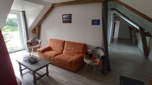 A cozy living room with a wooden ceiling, a sofa, two patterned chairs, a small table, and a view of a garden through a window.