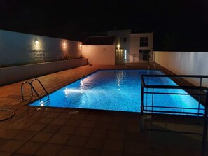 A rectangular swimming pool with a tiled deck, surrounded by a low wall and a small building in the background.