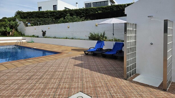 Una piscina con borde de baldosas, dos sillas reclinables con cubiertas azules y un edificio blanco de diseño moderno.