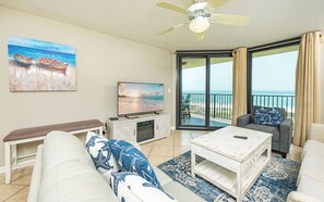 A living room with a TV, a bench, a sofa, a coffee table, and a view of the beach through large sliding doors.