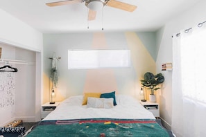 Une chambre à coucher avec un lit, un ventilateur de plafond, une fenêtre avec des stores, des tables de chevet avec des lampes et une plante.
