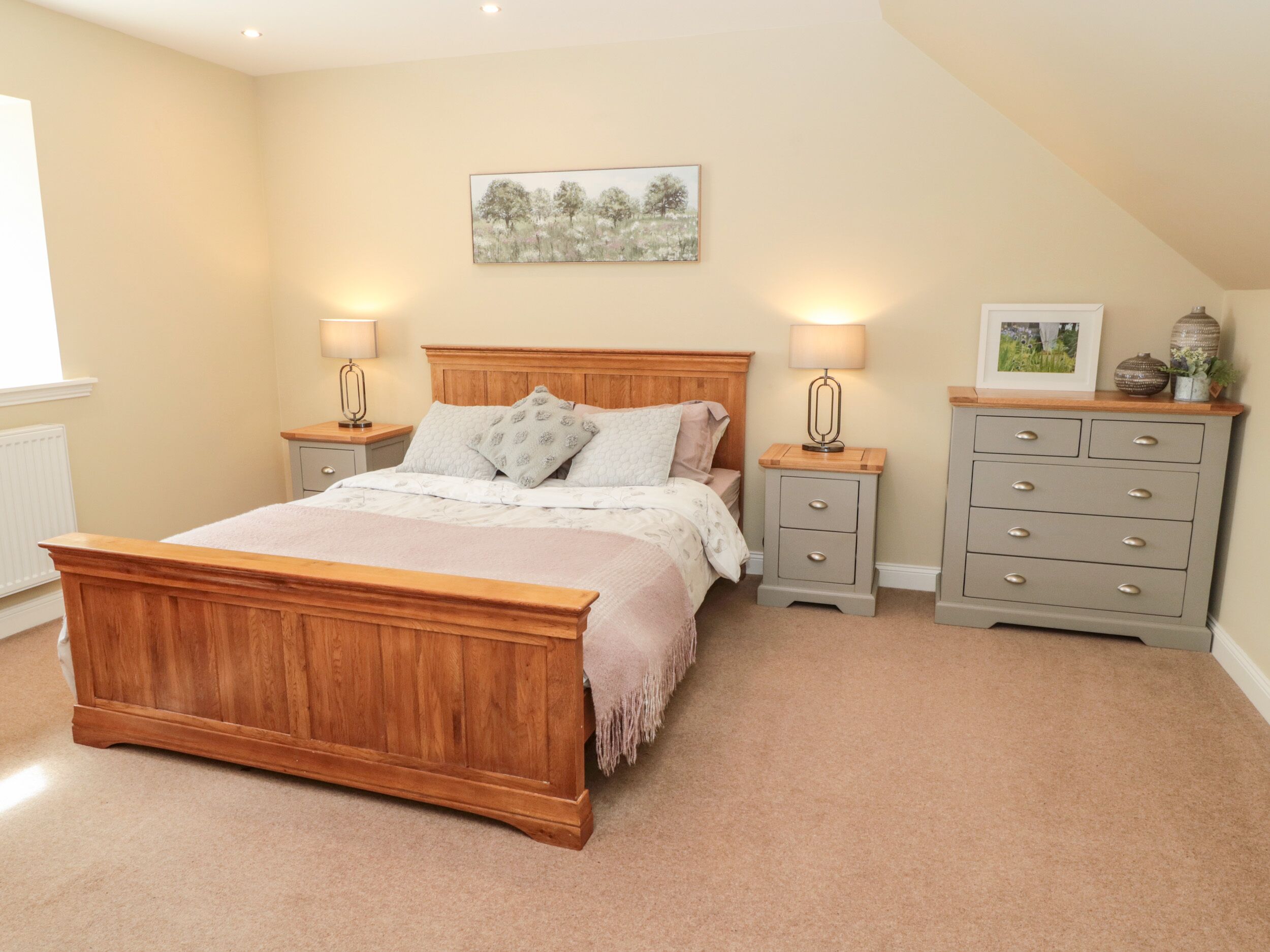 A bedroom with a wooden bed, two bedside tables with lamps, a grey dresser, and a wall-mounted picture.