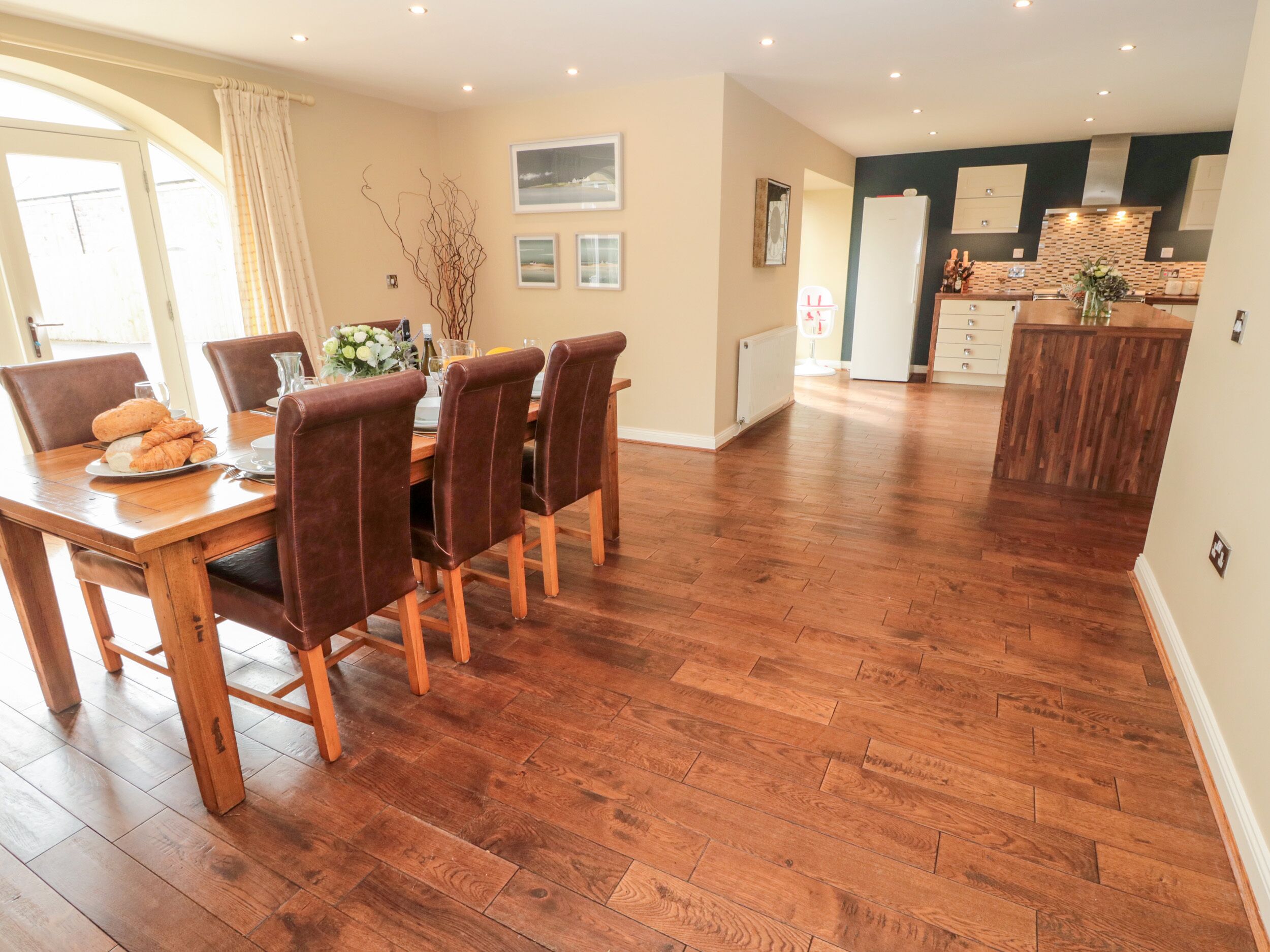 A dining area with a wooden table and chairs, a kitchen in the background, and a herringbone-patterned floor.