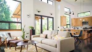 A spacious living area with a sofa, coffee table, and chairs, a kitchen with wooden cabinets, and large windows offering a view of greenery.