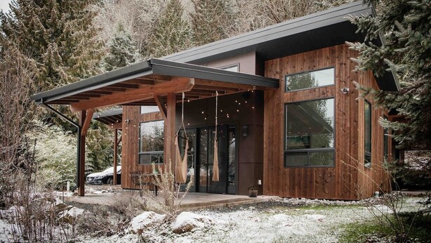 A modern wooden house with large windows, a covered porch, and a snowy landscape.