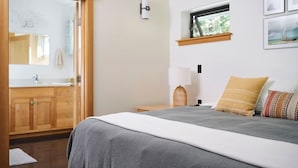 A bedroom with a bed, a nightstand, a lamp, a window, and a bathroom visible through an open door.
