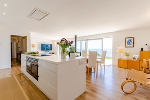 Cozinha moderna com ilha central, área de jantar com vista e sala de estar com banco de madeira.