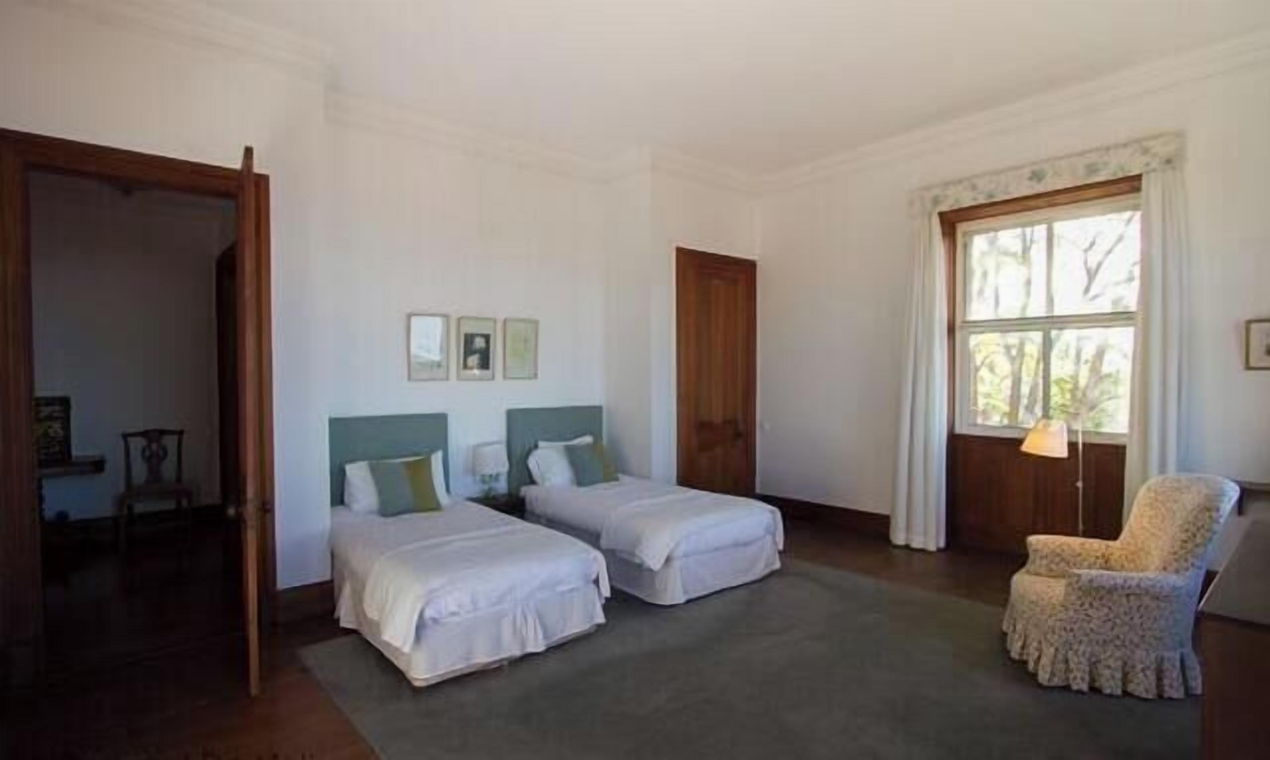 A bedroom with two single beds, a wooden door, a window with curtains, and a chair.