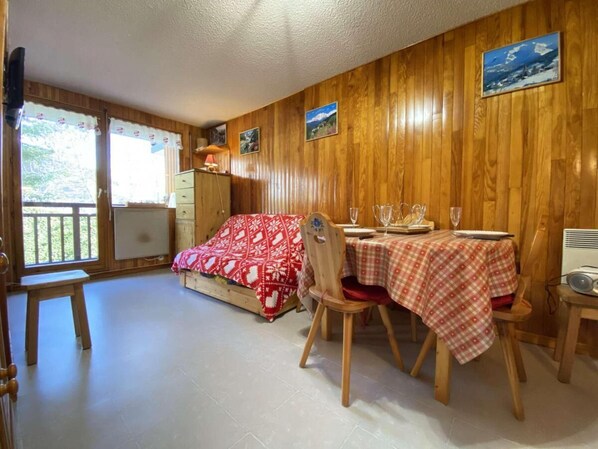 A cozy room with wooden walls, a bed with a red patterned blanket, a dining table with a checkered tablecloth, and a small table with glasses and a radio.