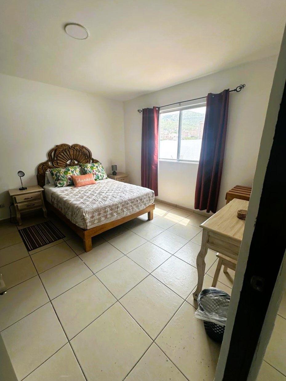 A bedroom with a bed, a bedside table, a nightstand, a window with curtains, and a tiled floor.