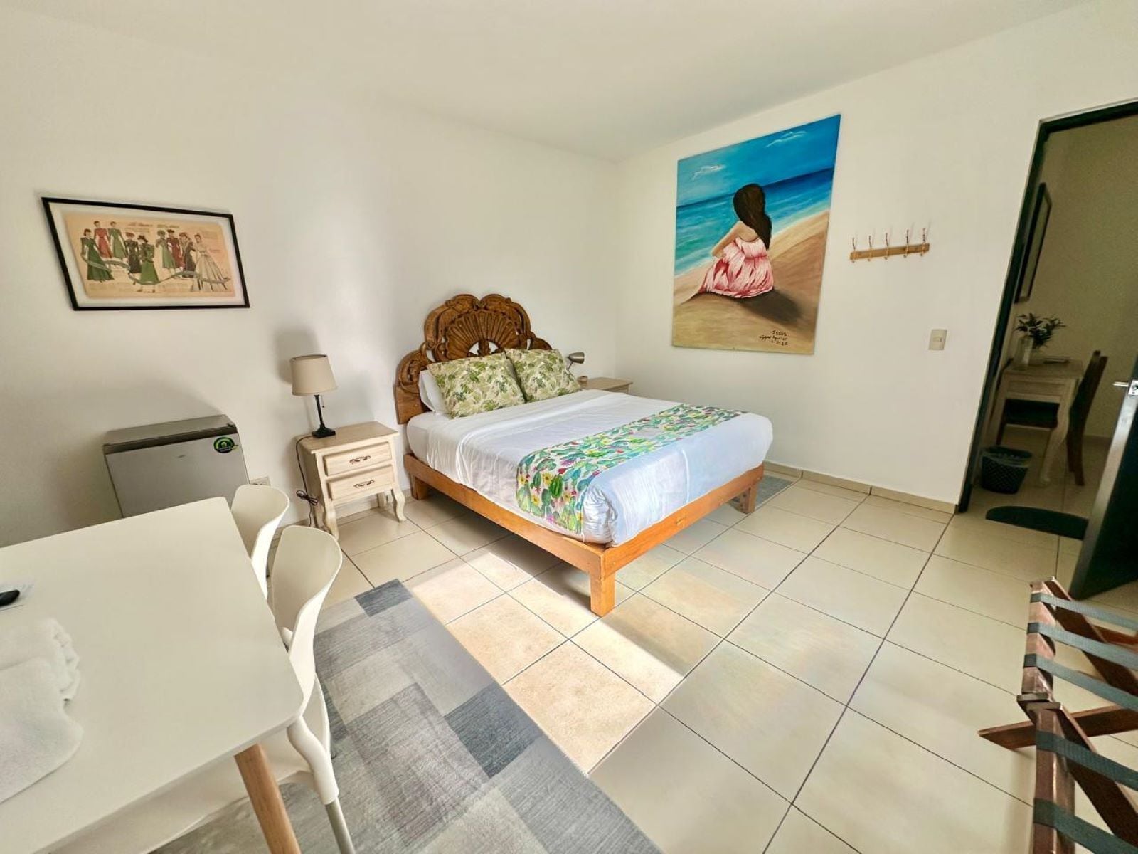 A bedroom with a bed, a desk, a chair, a nightstand, a lamp, a painting, and a small table with a plant.