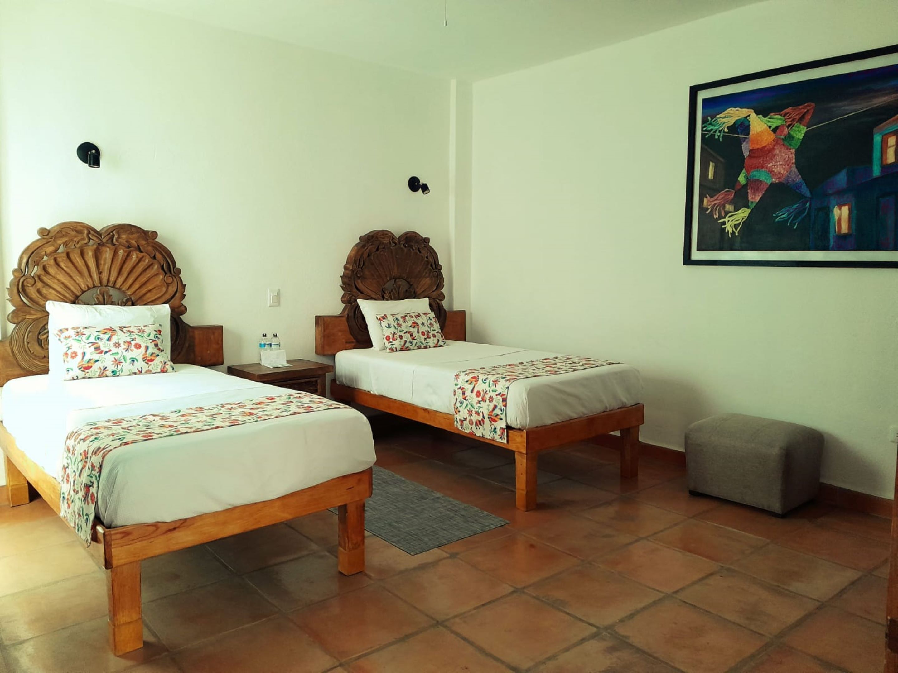 A hotel room with two wooden beds, a small gray ottoman, and a painting on the wall.