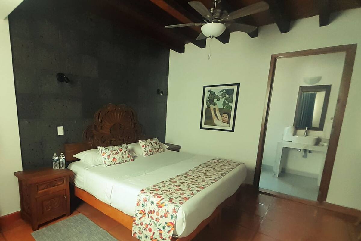 A bedroom with a bed, wooden nightstand, ceiling fan, and a bathroom visible through an open door.