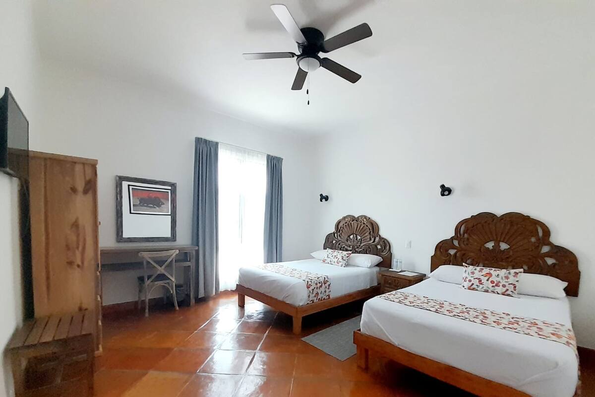 A room with two beds, a desk, a chair, a ceiling fan, and a framed picture on the wall.