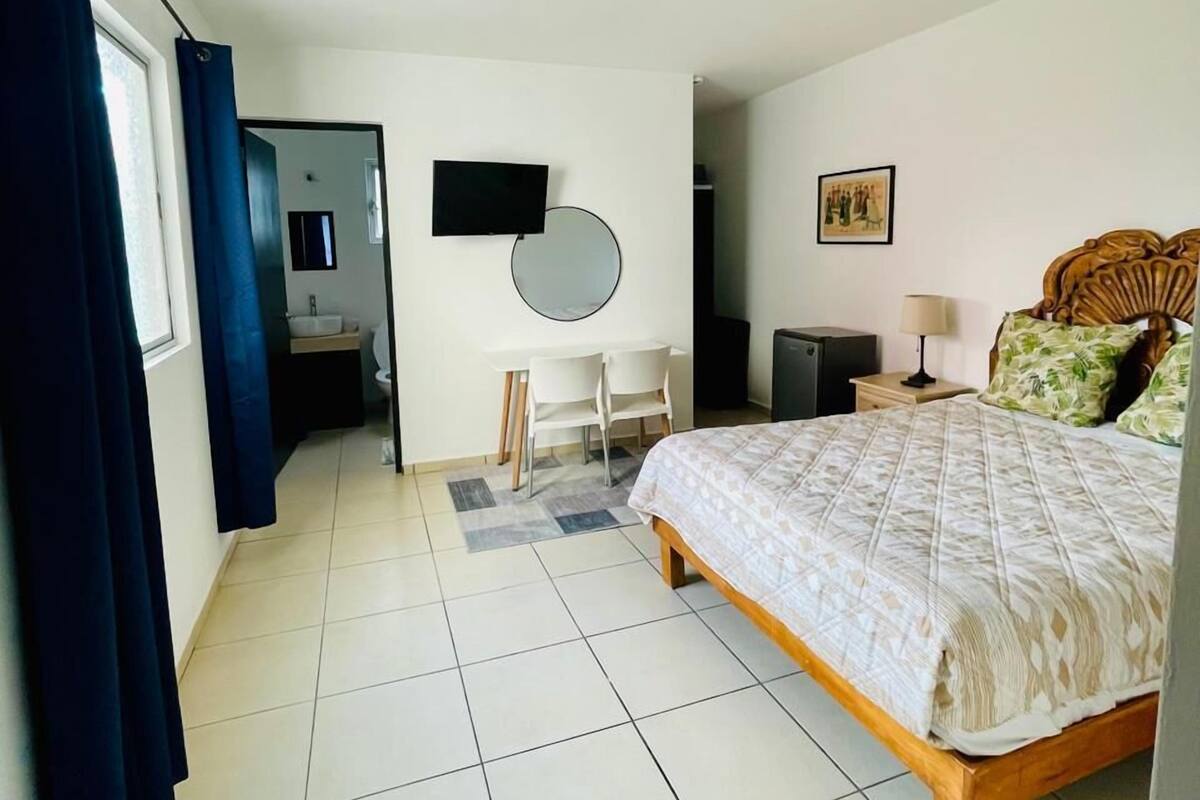 A bedroom with a bed, a desk, a chair, a TV, a mirror, and a nightstand.