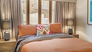 A hotel room with a large bed, two bedside lamps, and a view of buildings through the window. - Farrah - Designer, Three Bedroom Carlton Residence (Carlton)