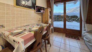 A dining area with a table set for four, wooden chairs, a television mounted on the wall, and a view of a snowy landscape through a glass door. - Ground floor, view mountain, terrace, tv, ski locker, 29m², Praz sur Arly (Praz-sur-Arly)