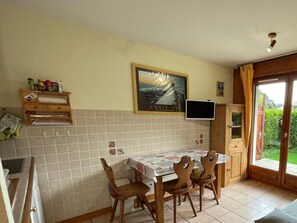 A kitchen with a tiled backsplash, wooden chairs, a table with a tablecloth, a painting on the wall, a television, and a sliding glass door leading to a garden. - Ground floor, view mountain, terrace, tv, ski locker, 29m², Praz sur Arly (Praz-sur-Arly)