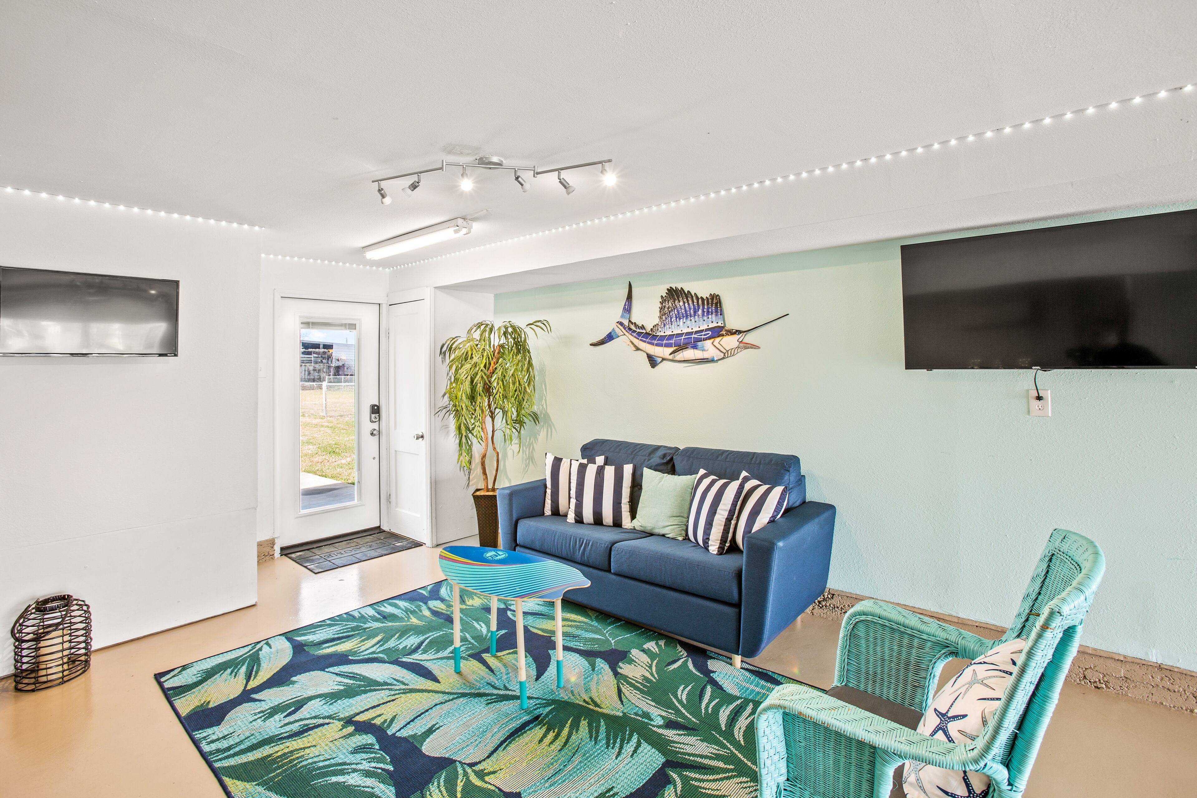 Uma sala de estar com um sofá azul, uma poltrona verde, um tapete estampado, uma planta em vaso e uma TV fixada na parede.