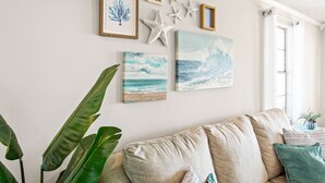 Una sala de estar con un sofá beige, una planta grande y decoración de pared que incluye cuadros y estrellas de mar.
