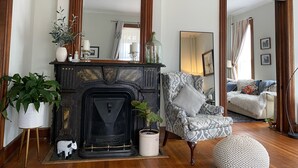 A living room with a fireplace, a patterned armchair, and a large mirror. - Historic Victorian on the Park (Little Falls)