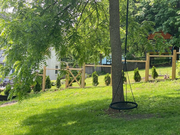 A tree with a swing hanging from it, surrounded by a grassy area with a wooden fence and a building in the background.