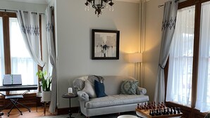 A living room with a sofa, a coffee table, a chessboard, a piano, and a chandelier. - Historic Victorian on the Park (Little Falls)