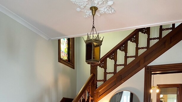 A grand staircase with intricate woodwork, a white armchair, a blue patterned rug, and a chandelier. - Historic Victorian on the Park (Little Falls)