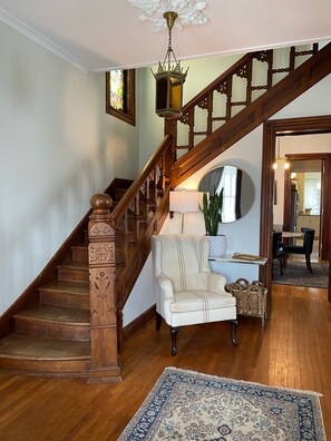 A grand staircase with intricate woodwork, a white armchair, a blue patterned rug, and a chandelier.