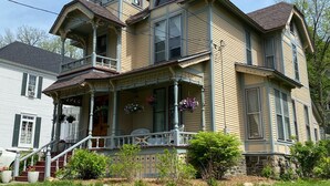 A large, two-story house with a prominent tower, a porch with white railings, and a stone foundation. - Historic Victorian on the Park (Little Falls)