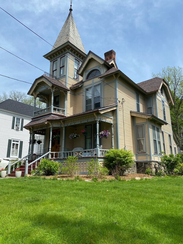 A large, two-story house with a prominent tower, a porch with white railings, and a stone foundation. - Historic Victorian on the Park (Little Falls)