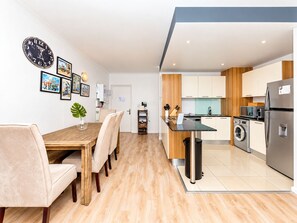 A modern kitchen with a built-in washing machine, a dining area with a wooden table and chairs, and a living space with a large clock and framed pictures on the wall. - Chic Beachfront Apt with Table Mountain views (Cape Town)