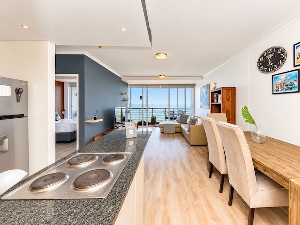 A modern kitchen with a central island, a dining area with a wooden table and chairs, and a living space with a sofa and a view of the sea.