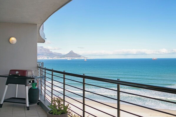 A balcony with a grill, a potted plant, and a view of the ocean and a mountain.